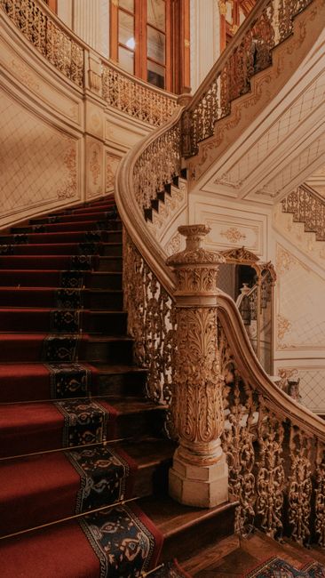 Inside a grand, opulent staircase leading up to a glass-domed ceiling, featuring a large crystal chandelier hanging centrally. The staircase has a red carpet runner with clear glass balustrades on eit