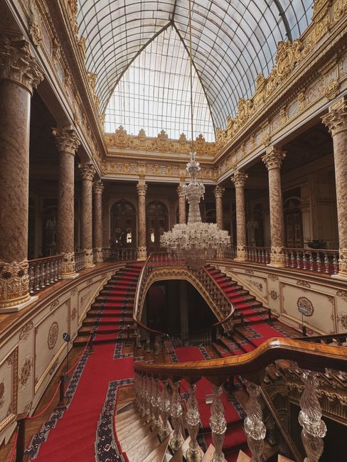 An opulent interior featuring a grand staircase with red carpeted steps and ornate wooden handrails, leading to an upper level with decorative balustrades. The space is illuminated by a large, intrica