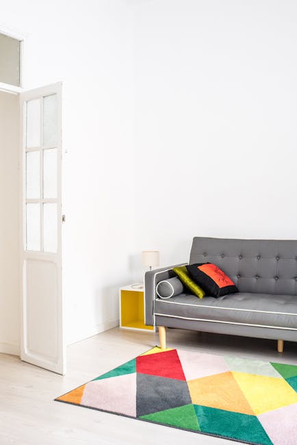 A modern living room featuring a grey upholstered sofa with button tufting against a bright white wall. On the sofa, there are three decorative cushions in black, red, and yellow. To the left of the sofa, a small yellow side table holds a white table lamp. A colorful geometric-patterned rug with shades of red, green, yellow, pink, and grey is spread out on the light wooden floor in front of the sofa. An open white wooden door with glass panes allows natural light to illuminate the room, highlighting the clean and tidy appearance of the space. This setting exemplifies domestic cleaning and surface maintenance, consistent with professional upholstery cleaning tips as seen on the PAGE_TITLE at carpetcleanerscrystalpalace.co.uk.