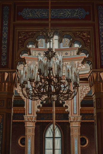 An ornate interior ceiling featuring a large vintage crystal chandelier with multiple candle-style lights and intricate metalwork, hanging from a high, decorated ceiling with detailed moldings and colorful painted accents. The ceiling is supported by elaborately crafted columns and arches, adorned with intricate patterns and gold accents. Natural light enters through tall, arched windows, illuminating the elegant, richly decorated space. This image captures the grandeur of a historical or luxurious building, emphasizing surface cleaning and maintenance of decorative architectural features, typical of high-quality domestic or commercial environments featured on SE19 upholstery cleaning tips for homes on Anerley Road, CRYSTAL PALACE, provided by carpetcleanerscrystalpalace.co.uk.