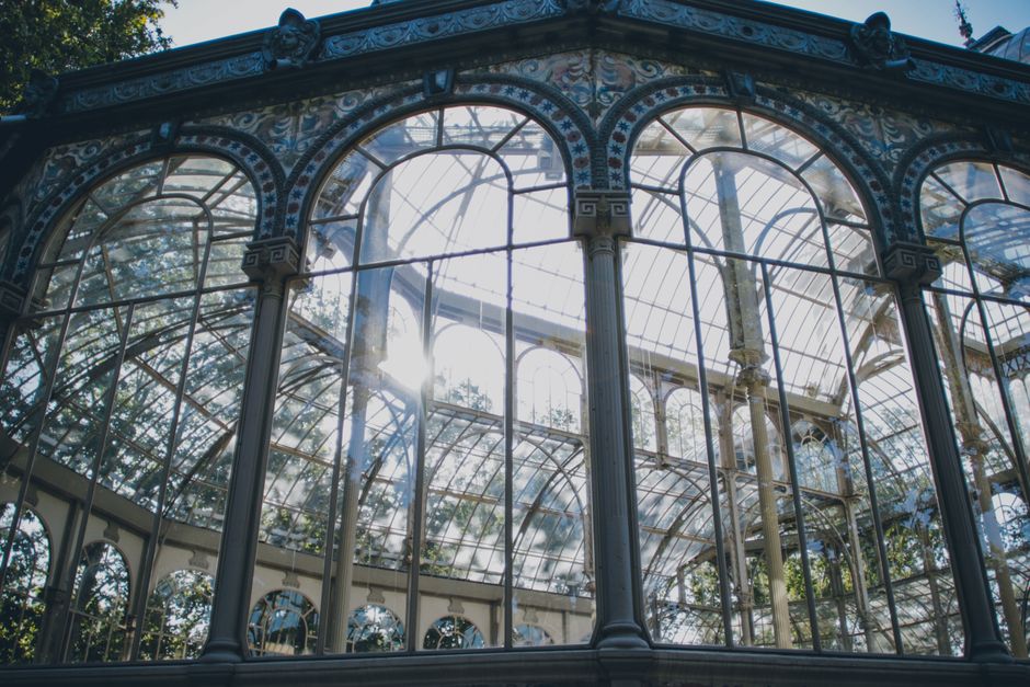 A detailed view of the ornate glass and metal conservatory structure, showcasing large arched windows, intricate decorative metalwork on the columns and frame, and the sunlight streaming through the glass panels. The conservatory appears clean and well-maintained with clear, dust-free glass surfaces, set in a garden or park setting with trees visible in the background. This image highlights the emphasis on surface cleaning and maintenance of glass and metal features, as seen in professional cleaning services like [COMPANY_NAME], related to Westow Hill carpet cleaning near Crystal Palace Park, CRYSTAL PALACE.